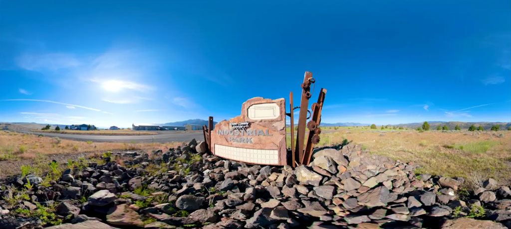 Weathered “INDUSTRIAL PARK” sign surrounded by dark volcanic rocks in a dry plain beneath a bright blue sky.