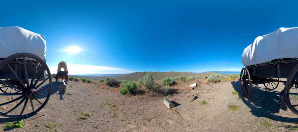 wo covered wooden wagons frame a dry, shrub-covered landscape with low hills beneath a bright blue sky.