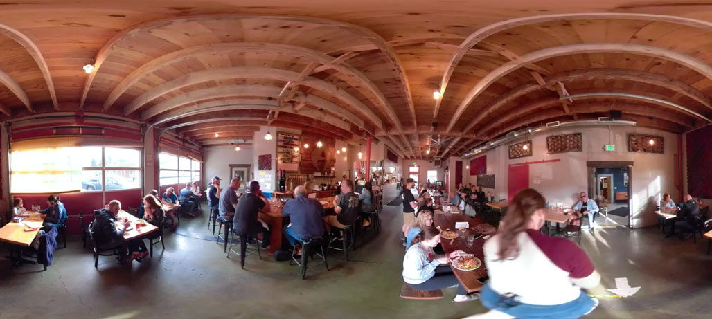 Panoramic view of a busy restaurant with exposed wooden beams, red walls, communal seating, and diners throughout the room.