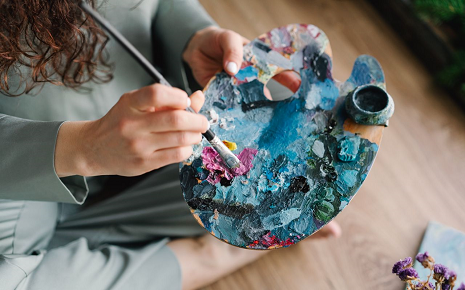 The image shows a close-up of a person holding a wooden artist's palette covered in colorful paint while using a brush.
