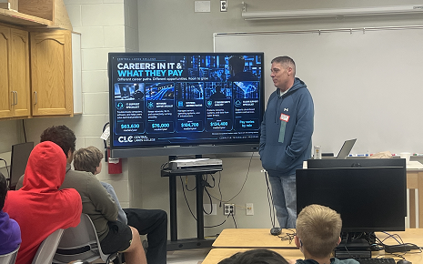 The image shows a presenter speaking to a classroom of students about information technology career paths and salaries displayed on a large screen.