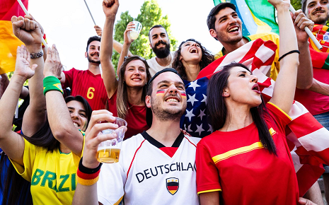 The image is a stock photo showing a multi-ethnic group of soccer fans in bright soccer shirts cheering together.