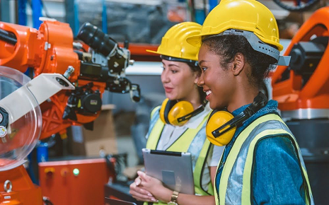 The image shows two women wearing safety helmets, high-visibility vests, and ear protection as they inspect or operate an industrial robotic arm in a modern manufacturing facility.