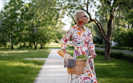 This image shows an elegant senior woman with short grey hair and large red earrings wearing a colorful floral-patterned dress while walking beside a bicycle with a basket of groceries in a park.