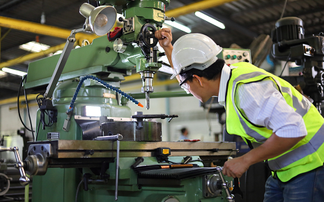 This is an image of an industrial worker in a hard hat and safety vest operates a precision milling or machining tool inside a factory.
