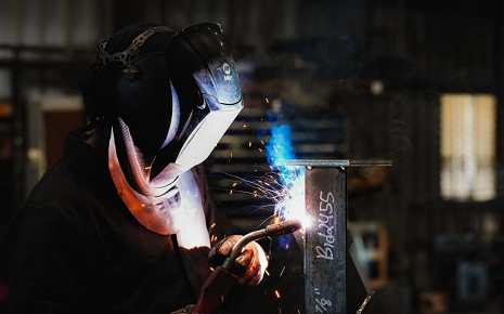 The image shows a worker wearing a dark protective suit, thick work gloves, and an auto-darkening welding helmet while performing metal arc welding on a vertical steel piece.