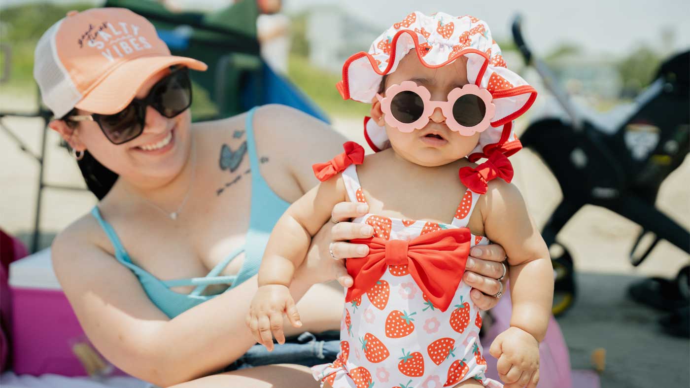 Mama holding her baby who is wearing a strawberry swimsuit and pink flower-shaped sunglasses at El Jardin Beach in Pasadena, TX