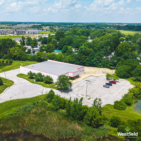 The image shows an aerial view of a single-story commercial/retail building with a flat roof, red trim accents, an adjacent asphalt parking lot, and surrounding greenery next to a body of water or wetland area.