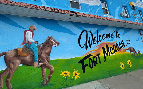The image shows a large outdoor mural painted on a building in Fort Morgan, Colorado, featuring the text "Welcome to Fort Morgan, CO" alongside a cowboy on horseback, a windmill, and sunflowers.