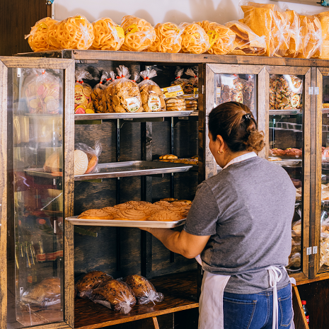The image shows a bakery employee stocking fresh shelves with traditional pan dulce (Mexican sweet bread) from a baking sheet.