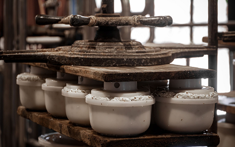 The image shows a mechanical screw press used to shape or press ceramic or pottery items in a workshop. Rows of white ceramic-like forms or molds sit on wooden shelves beneath a hand-operated metal and wood pressing mechanism.