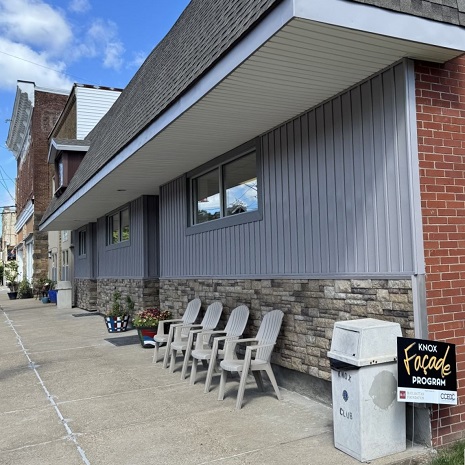 The image shows a commercial building exterior on Main Street in Knox, Pennsylvania, featuring gray vertical siding, a stone veneer base, a wide overhang roof, and a sign for the Knox Façade Program.
