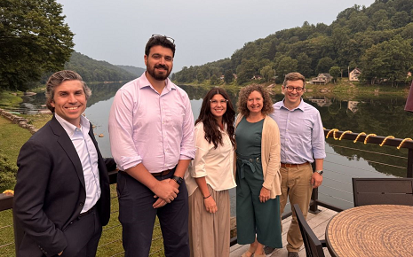 This image shows a group of people standing on a wooden deck or patio overlooking a river framed by forested hills, a scenic riverfront location.