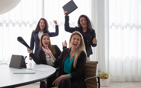 This image features four women in business attire, two seated and two standing cheering enthusiastically in an office setting with sheer curtained windows in the background.