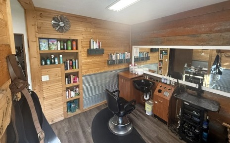 Interior of a compact hair salon featuring warm wood-paneled walls, a styling station with a large mirror, a shampoo sink, a rolling utility cart, and shelving stocked with products.