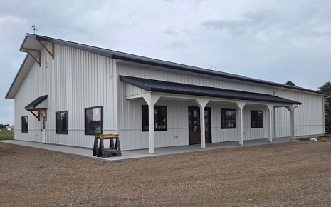 The image shows a white metal pole-frame building or barndominium with a dark metal roof, a front-facing gable, and an attached covered porch supported by white posts.