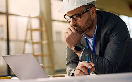 A man in a hard hat leaning over a table and looking at information on a laptop