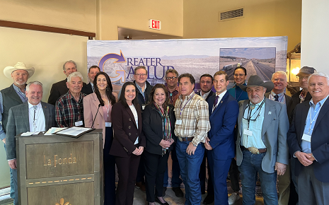 This is an image of eighteen Economic Roundtable Participants staning in two rows , with a podium on the left, foreground. In the background is a large board with the Greater Gallup logo.