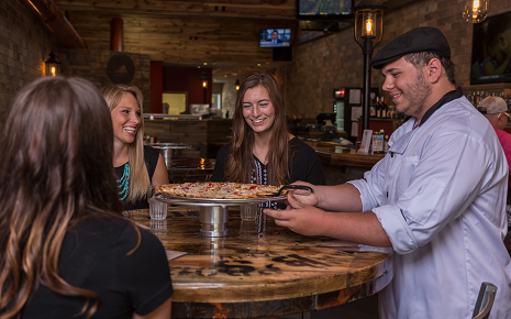 This image shows a group of people dining at a restaurant or tavern with a chef serving a freshly baked pizza on a wooden peel.
