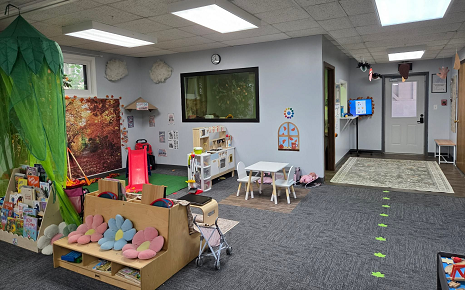 The image shows an indoor preschool or daycare classroom equipped with children's play furniture, toy storage shelves, a small table and chairs, a play kitchen, a green canopy tree structure, and grey carpeting.